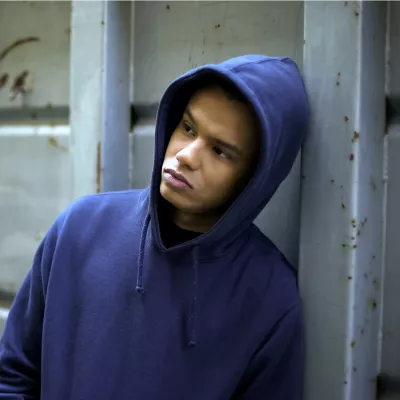 Young mixed race person in blue hoodie leaning against concrete wall